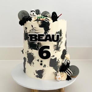 May include: A birthday cake with white frosting and black brushstrokes, decorated with Star Wars-themed figurines and cookies. The name "BEAU" and the number "6" are displayed on the cake. The cake sits on a white cake stand.