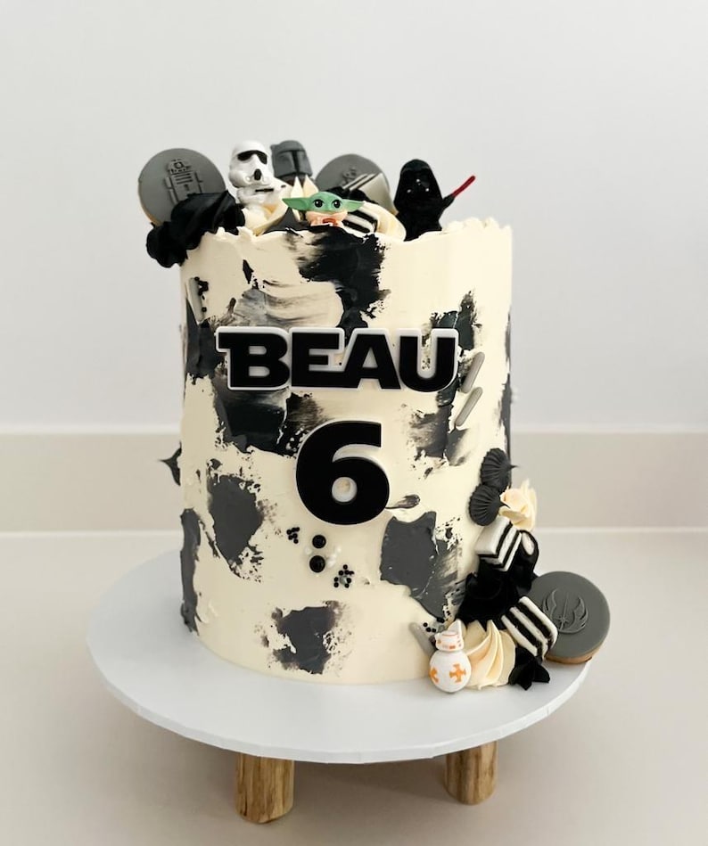 May include: A birthday cake with white frosting and black brushstrokes, decorated with Star Wars-themed figurines and cookies. The name "BEAU" and the number "6" are displayed on the cake. The cake sits on a white cake stand.
