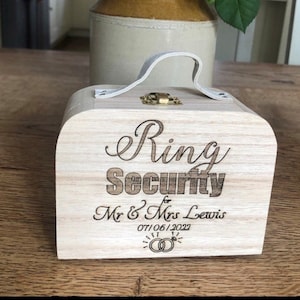 May include: A wooden ring box with a white handle and a brass latch. The box is engraved with the text "Ring Security for Mr. & Mrs. Lewis 07/06/2022" and two interlocking rings.