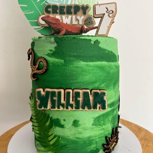 May include: A green frosted cake with a jungle theme. The cake is decorated with wooden accents, including a lizard, the number 7, and the name "William". The cake also features a "Creepy Crawly" topper and green leaf decorations.