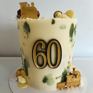 May include: A white cake decorated with gold and green accents. The cake has a large gold number 60 on the front and a gold train on the bottom.