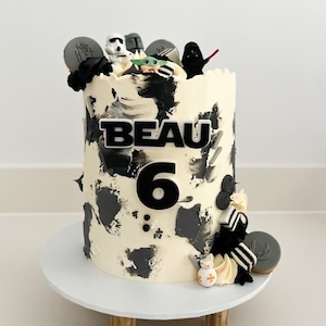May include: A birthday cake with white frosting and black brushstrokes, decorated with Star Wars-themed figurines and cookies. The cake has the name "BEAU" and the number "6" on it. It sits on a white cake stand.
