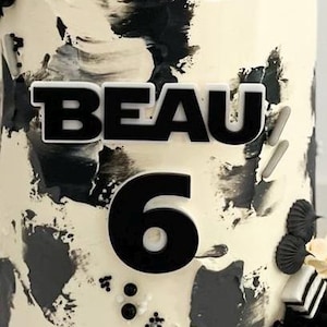 May include: A decorated cake with a white base and black abstract brushstrokes. The word "BEAU" and the number "6" are prominently displayed in black. The cake is adorned with decorative elements.