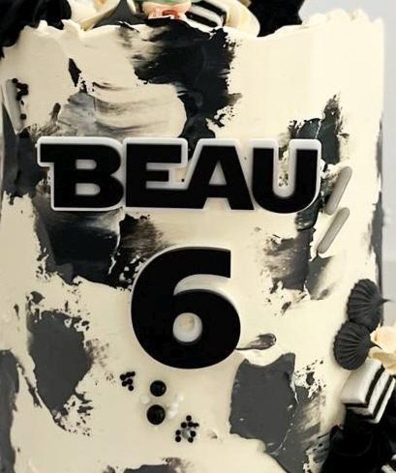 May include: A decorated cake with a white base and black abstract brushstrokes. The word "BEAU" and the number "6" are prominently displayed in black. The cake is adorned with decorative elements.
