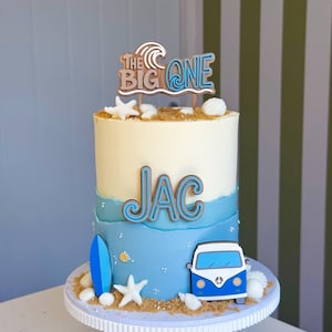 May include: A tiered cake with white and blue frosting, decorated with edible sand, white starfish, and seashells. It features a blue surfboard, a blue and white van, and wooden toppers reading "THE BIG ONE" and the name "JAC" in blue and brown.