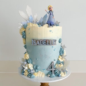 May include: A blue and white frosted cake decorated with a Frozen character, snowflakes, and a silver 'Isabelle' name topper. The cake is decorated with a number '4' on the side.