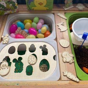 May include: A wooden play table with two white containers filled with colourful plastic eggs. The table also has a green container with brown material and a white bowl. There are various wooden cutouts of animals and vegetables on the table.
