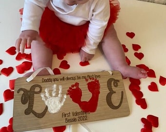 Può includere: Un cartello di legno con la parola "LOVE" scritta in corsivo. Le lettere sono decorate con un'impronta di mano bianca e due impronte di piedi rosse. Il cartello include anche il testo "Daddy you will always be my first love. First Valentine's Bessie 2022."