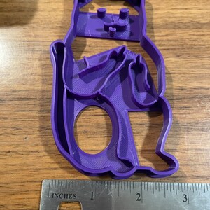 K&ouml;nnte beinhalten: Lilafarbener Pl&auml;tzchenausstecher aus Kunststoff in Form einer sitzenden Katze mit verschr&auml;nkten Armen und ausgestrecktem Mittelfinger. Der Ausstecher ist etwa 7,5 cm hoch.