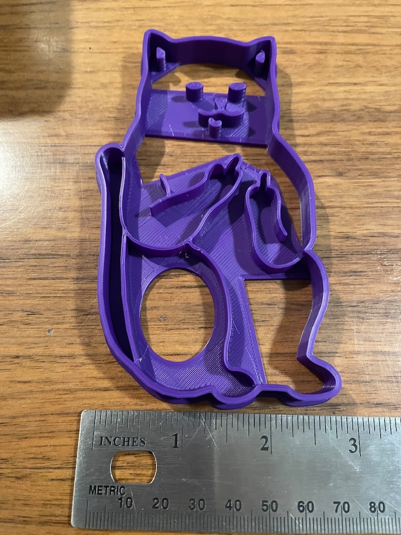 K&ouml;nnte beinhalten: Lilafarbener Pl&auml;tzchenausstecher aus Kunststoff in Form einer sitzenden Katze mit verschr&auml;nkten Armen und ausgestrecktem Mittelfinger. Der Ausstecher ist etwa 7,5 cm hoch.