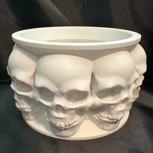 May include: White ceramic planter shaped like a circle of skulls. The planter has a smooth, white finish and is perfect for displaying plants or flowers.
