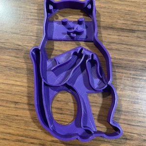 K&ouml;nnte beinhalten: Ein lilafarbener Pl&auml;tzchenausstecher aus Kunststoff in Form einer Katze, die mit verschr&auml;nkten Armen sitzt und den Mittelfinger zeigt.