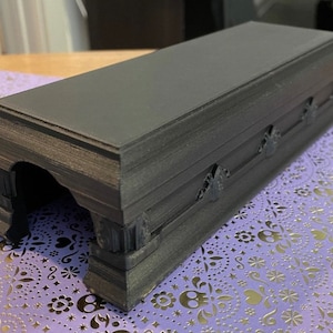 May include: A black coffin-shaped box with a textured surface and a small opening on one side. The box is sitting on a purple and silver patterned surface.