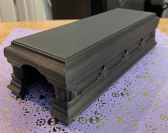 May include: A black coffin-shaped box with a textured surface and a small opening on one side. The box is sitting on a purple and silver patterned surface.