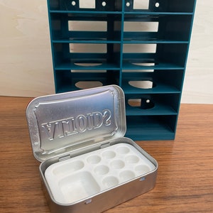 May include: A teal plastic storage organizer with multiple compartments, next to an open metal tin with the word "VIVID2" on the lid. The tin contains a white plastic tray with nine round wells for paint.