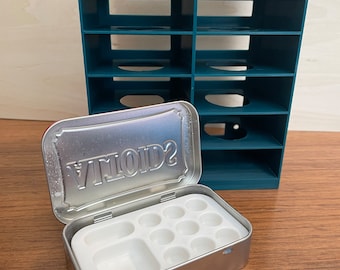 May include: A teal plastic storage organiser with multiple compartments, next to an open metal tin with the word "VIVID2" on the lid. The tin contains a white plastic tray with nine round wells for paint.