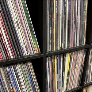 May include: A collection of vinyl records stored in a black wooden shelf. The records are in various colors and designs, with visible album art and titles. The shelf is divided into multiple sections, each filled with records.