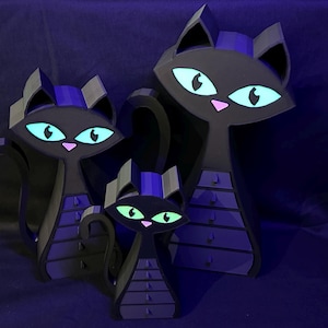 May include: Three stylized black cat figurines with glowing teal eyes and pink noses. The cats have a geometric design with a purple accent on their heads. The cats are arranged in a family grouping, with a large, medium, and small cat.