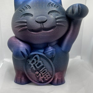 May include: A blue, purple, and black Maneki Neko cat figurine with a smiling face and raised paw. The figurine has a speckled finish and a coin with Japanese characters.