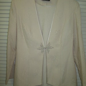 May include: A cream-colored suit jacket with a matching skirt. The jacket has a V-neckline and a decorative button closure. The label on the jacket reads "Ursula".