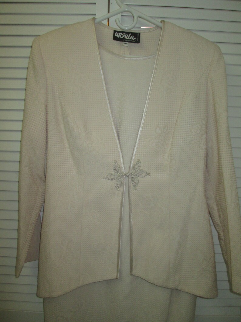 May include: A cream-colored suit jacket with a matching skirt. The jacket has a V-neckline and a decorative button closure. The label on the jacket reads "Ursula".