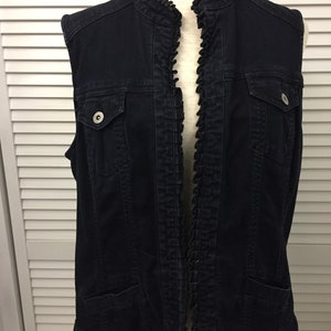 May include: A black denim vest with a ruffled collar and a zipper closure. The vest has two pockets on the front and a decorative ruffle trim around the edges.