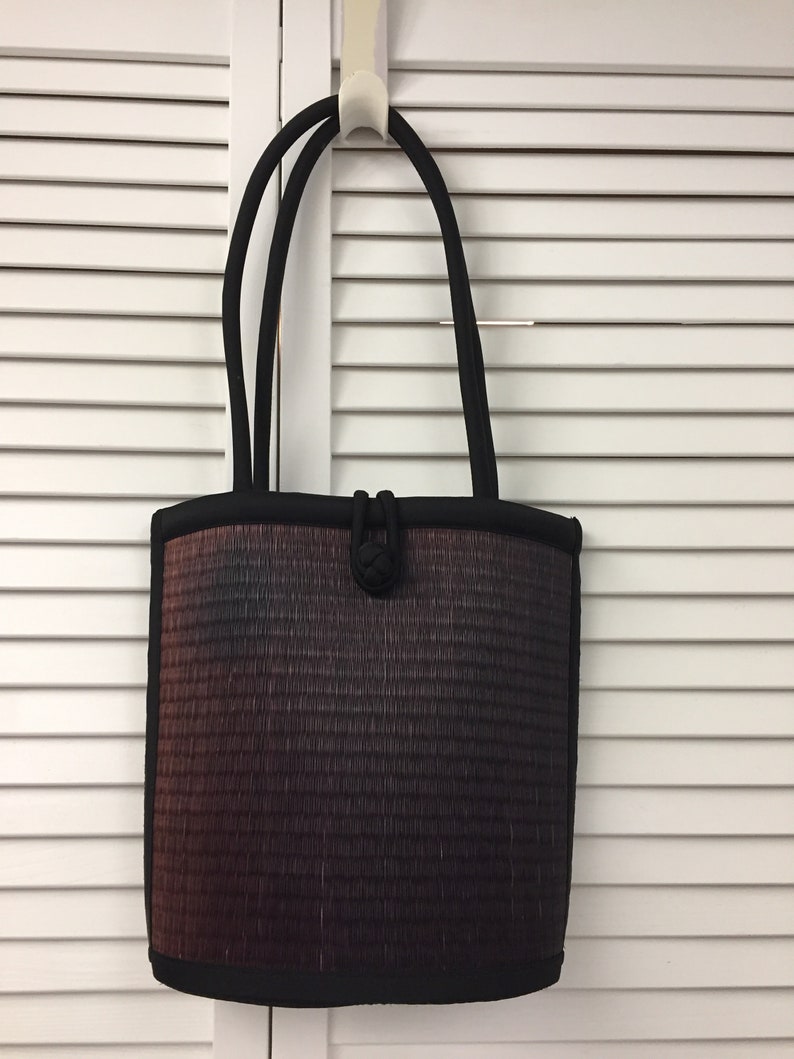 May include: A black tote bag with a woven design in shades of brown, gray, and black. The bag has two black handles and a knotted detail on the front.