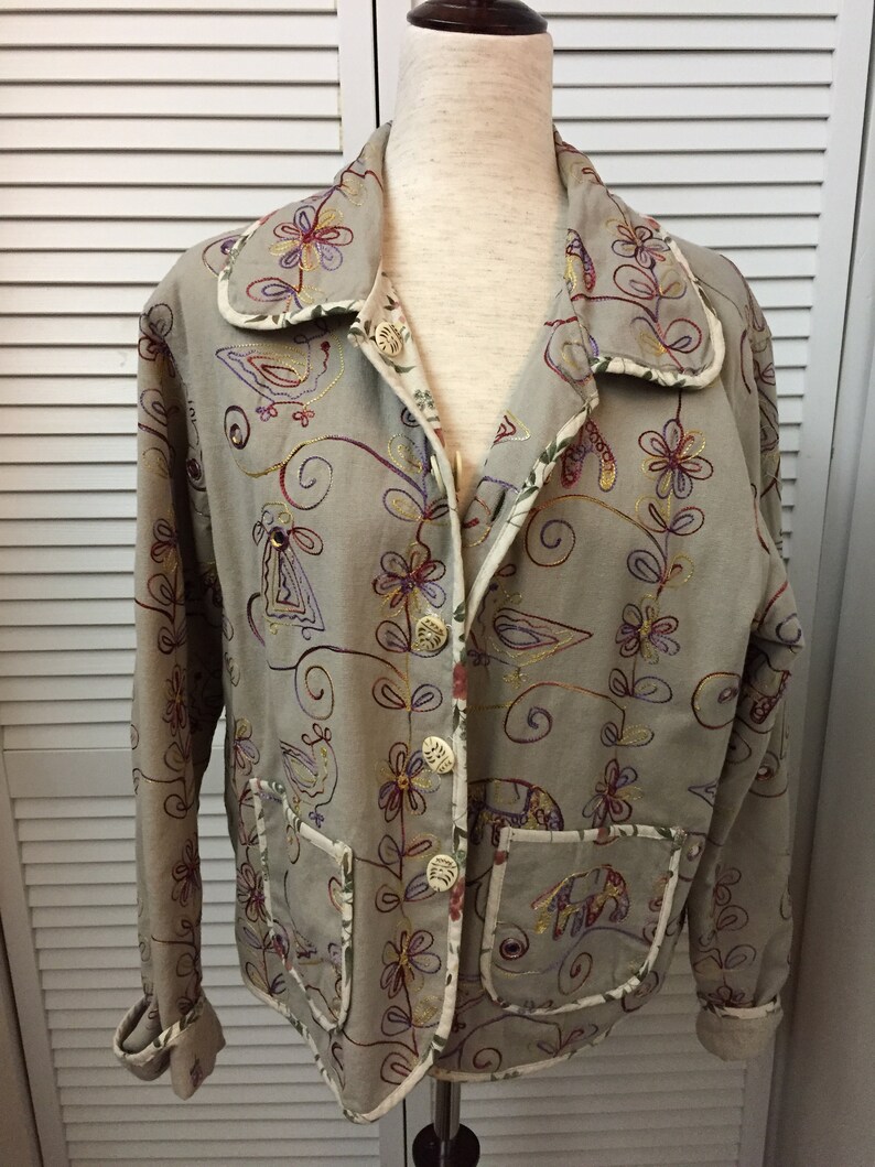 May include: A beige embroidered jacket with a floral and bird design. The jacket has a button closure and two pockets.