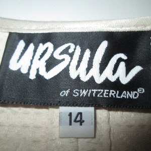 May include: A black label with white text that reads "Ursula of Switzerland" and "Made in USA". The label is attached to a white fabric garment with a white tag that reads "14".