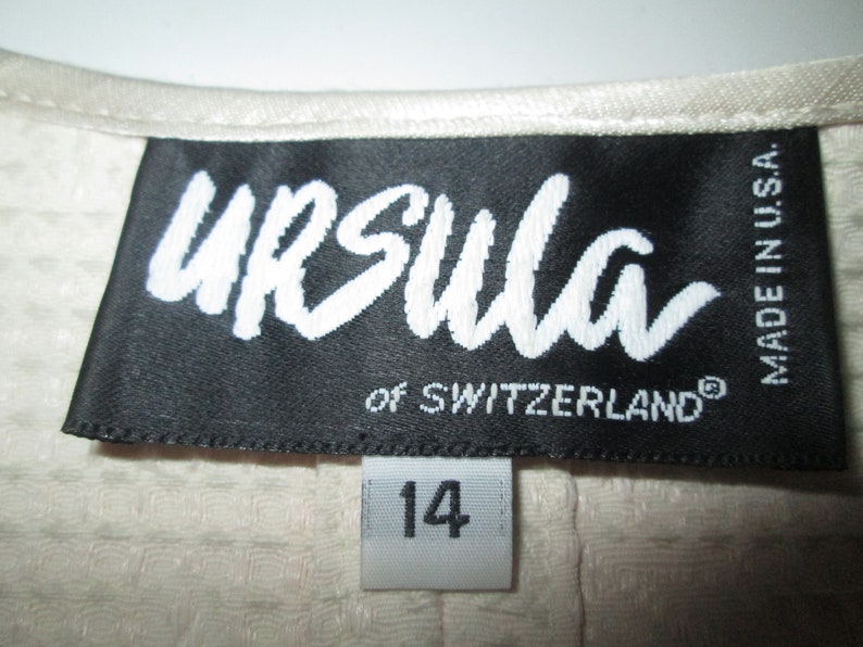 May include: A black label with white text that reads "Ursula of Switzerland" and "Made in USA". The label is attached to a white fabric garment with a white tag that reads "14".