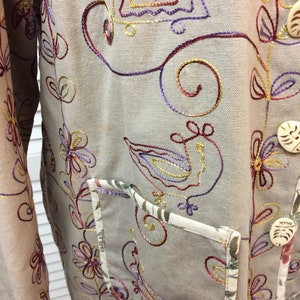 May include: A beige jacket with colourful embroidered floral designs. The jacket has a pocket on the front and two buttons.