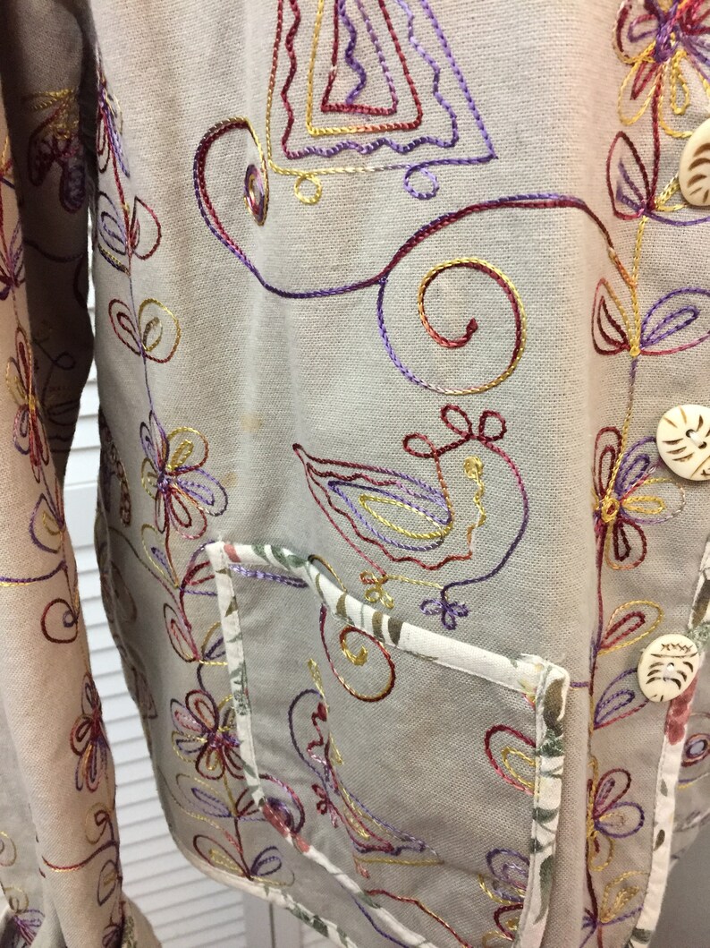 May include: A beige jacket with colourful embroidered floral designs. The jacket has a pocket on the front and two buttons.
