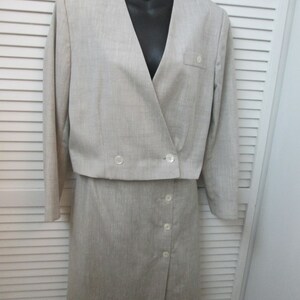 May include: A light beige two-piece suit consisting of a cropped long-sleeve jacket with a V-neckline and two front buttons, and a matching skirt featuring a side-button closure. The fabric has a subtle textured finish throughout the garment.