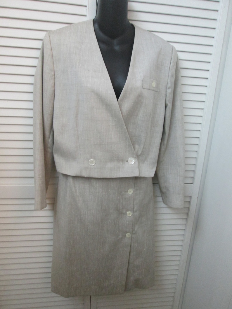 May include: A light beige two-piece suit consisting of a cropped long-sleeve jacket with a V-neckline and two front buttons, and a matching skirt featuring a side-button closure. The fabric has a subtle textured finish throughout the garment.