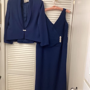 May include: A navy blue two-piece formal outfit. The set includes a blazer with a single button closure and a sleeveless dress with a draped bodice and a decorative embellishment. The dress hangs on a pink hanger.