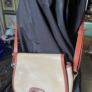 Puede incluir: Bolso bandolera beige y marrón con cierre de solapa. El bolso tiene un cuerpo beige con ribetes marrones y una correa de cuero marrón. Cuenta con una hebilla de metal dorada y un cierre de anilla en D. El bolso se lleva sobre una camisa negra.
