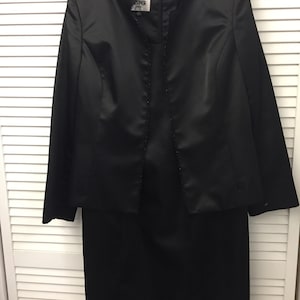 Op de afbeelding: Een zwart satijnen jurkenpak met een jasje en rok. Het jasje heeft een parelrand rond de halslijn en de voorste opening. Het label op het jasje staat "Kasper".