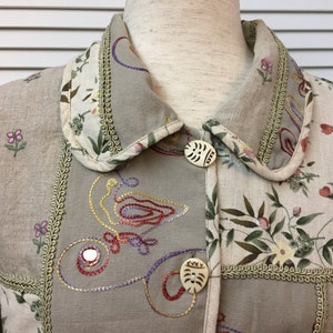 May include: A beige linen jacket with a floral print and embroidered details. The jacket has a collar with a decorative trim and two buttons.