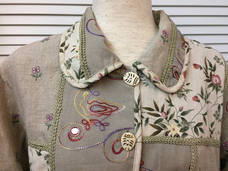 May include: A beige linen jacket with a floral print and embroidered details. The jacket has a collar with a decorative trim and two buttons.