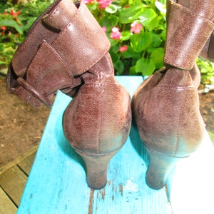 May include: A pair of brown leather ankle boots with a buckle detail. The boots have a low heel and are sitting on a blue surface.