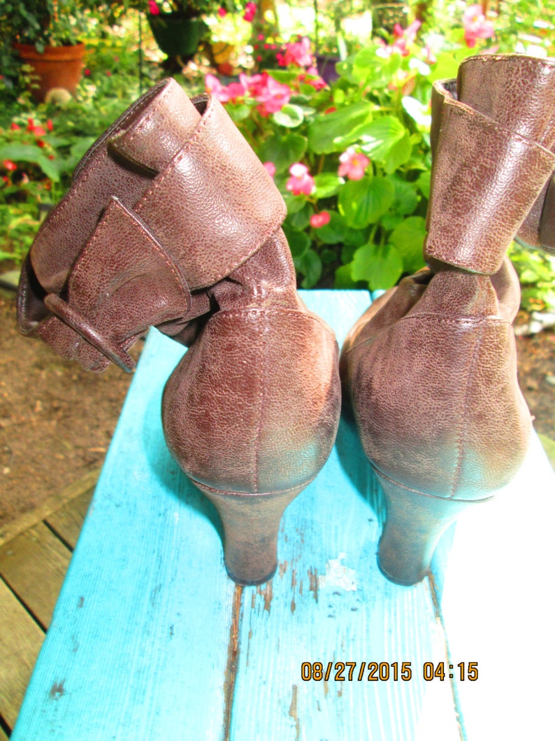 May include: A pair of brown leather ankle boots with a buckle detail. The boots have a low heel and are sitting on a blue surface.