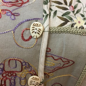 May include: A beige fabric with embroidered floral designs and two decorative buttons. The buttons are made of a light brown material and have a carved design.