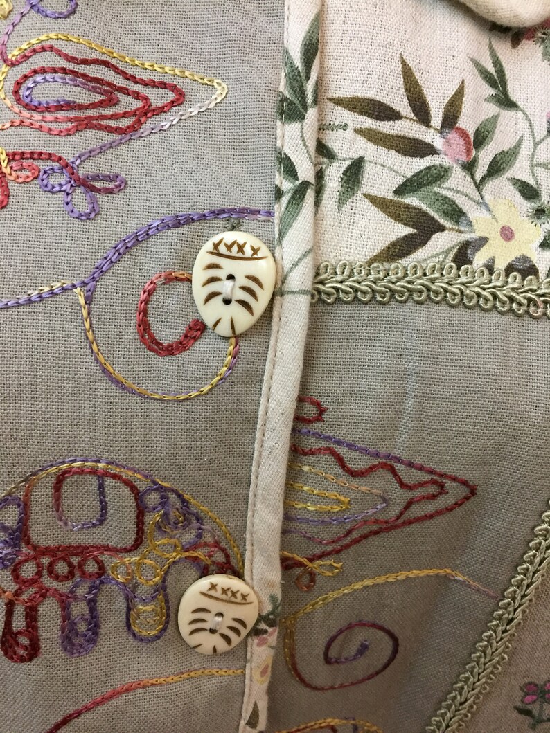 May include: A beige fabric with embroidered floral designs and two decorative buttons. The buttons are made of a light brown material and have a carved design.