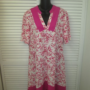 Può includere: Un abito bianco con stampa floreale rosa e un colletto a V rosa. L'abito ha maniche corte e una forma leggermente a trapezio.