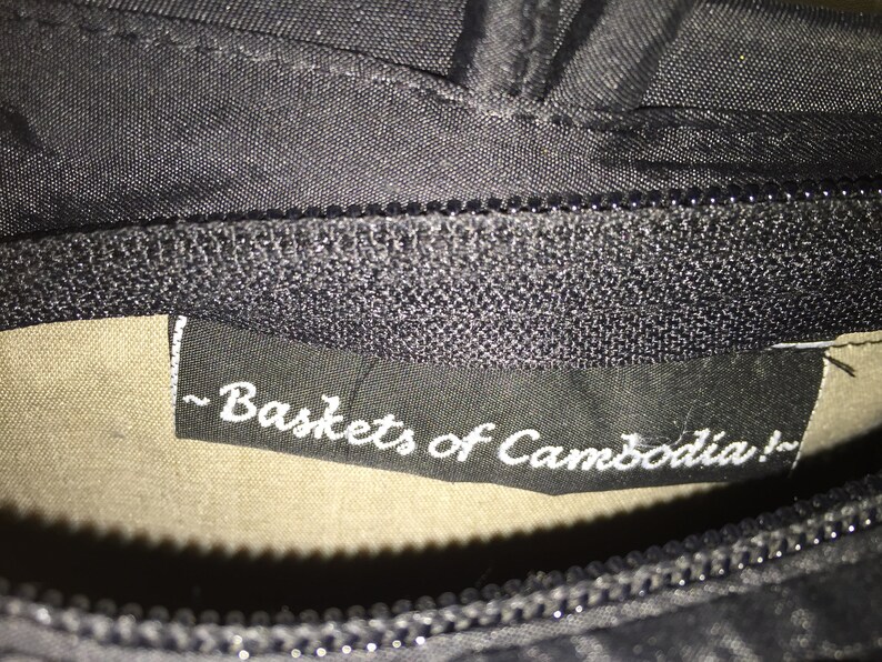 May include: Close-up of a black fabric label with white text that reads "~ Baskets of Cambodia! ~"