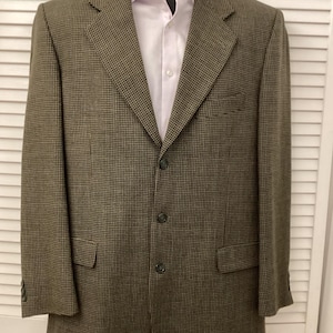 Könnte beinhalten: Ein braun-schwarzer Blazer mit Hahnentrittmuster und einem hellrosa Hemd darunter. Der Blazer hat drei Knöpfe und zwei Vordertaschen. Der Blazer wird auf einem schwarzen Schaufensterkopf präsentiert.