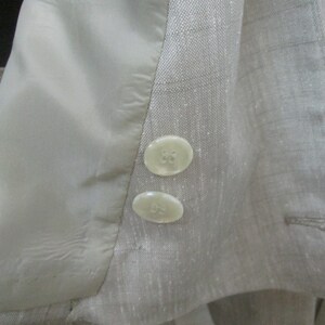 May include: A light beige blazer with a subtle grid pattern, featuring two round, off-white buttons on the sleeve cuff and a smooth, matching beige interior lining. The fabric has a visible, slightly textured weave throughout the garment.