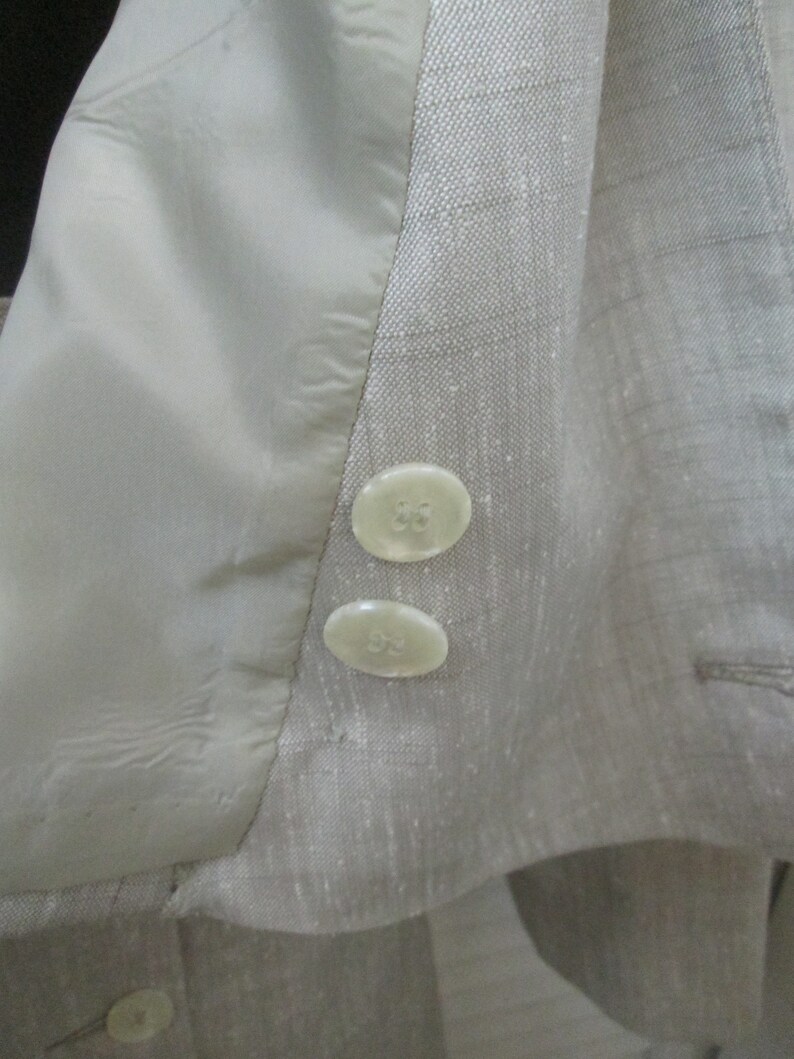 May include: A light beige blazer with a subtle grid pattern, featuring two round, off-white buttons on the sleeve cuff and a smooth, matching beige interior lining. The fabric has a visible, slightly textured weave throughout the garment.
