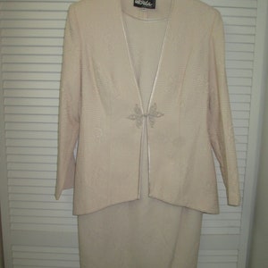 May include: A cream-colored suit jacket and skirt set with a delicate floral pattern. The jacket has a V-neckline and a small, decorative brooch in the center. The label "Ursula" is visible on the jacket.