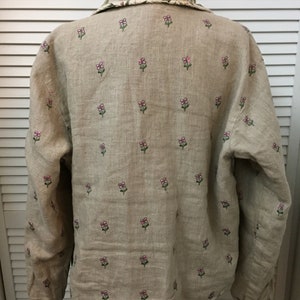 May include: A beige linen shirt with long sleeves and a floral pattern. The flowers are embroidered in pink and green.